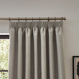 Dawn pencil pleat curtains in linen, showing pinch-pleated heading on a metal pole above a window in a neutral-toned room.