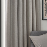 Dawn pencil pleat curtains in linen, shown in soft folds beside a grey armchair, with a woven marl-effect fabric in a muted beige tone.