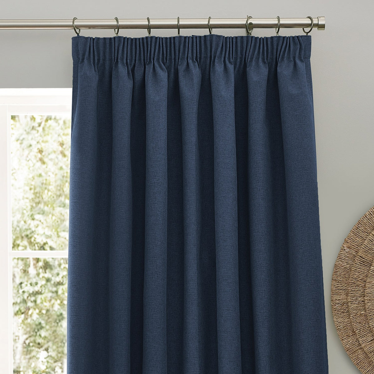 Dawn Plain Polyester Pencil Pleat Curtains in midnight blue, gathered on tape heading with metal rings on a pole, shown against a light grey wall near a window.