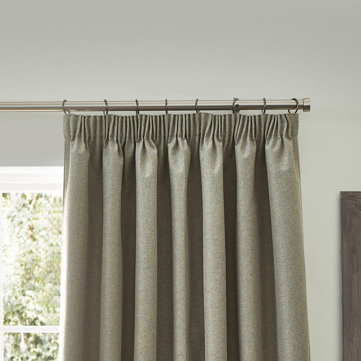 Dawn pencil pleat curtains in natural marl-effect polyester, shown gathered on rings along a metal pole beside a window in a light-toned room.