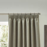 Dawn pencil pleat curtains in natural marl-effect polyester, shown gathered on rings along a metal pole beside a window in a light-toned room.