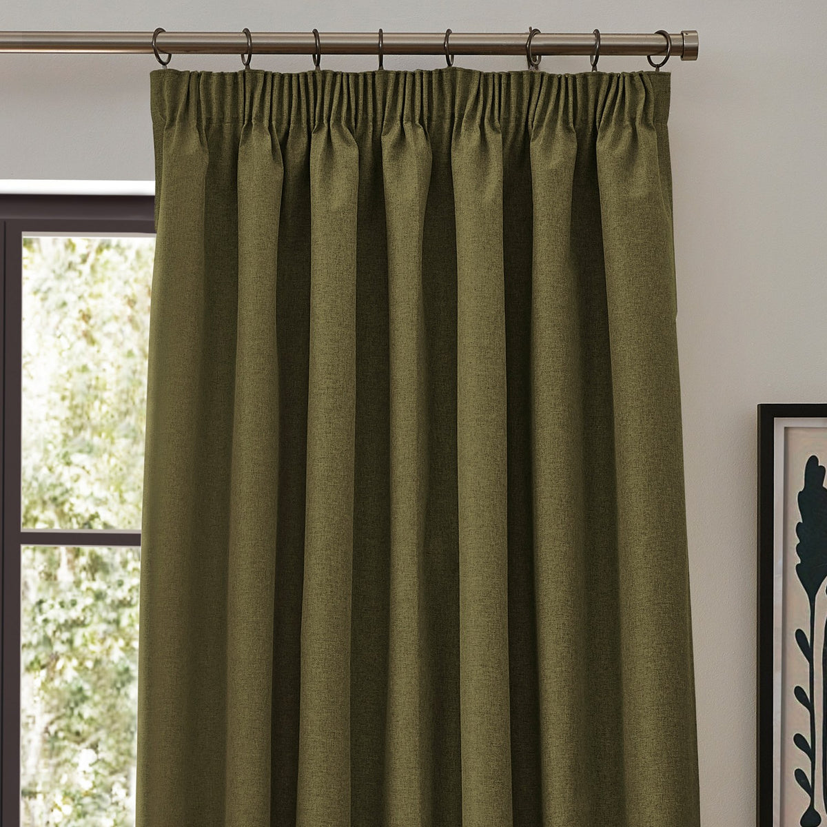 Dawn plain polyester pencil pleat curtain in olive green with pencil pleat header and metal rings, shown hanging on a pole beside a window in a living room.