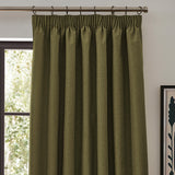 Dawn plain polyester pencil pleat curtain in olive green with pencil pleat header and metal rings, shown hanging on a pole beside a window in a living room.
