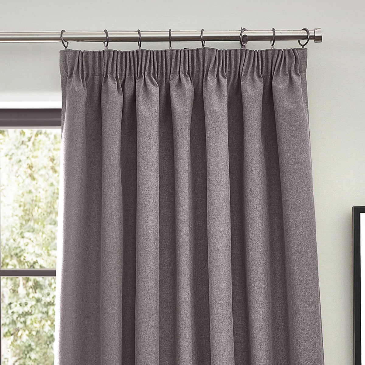 Dawn pencil pleat curtains in charcoal grey polyester with gathered heading tape, hung on curtain rings beside a window in a living room setting.