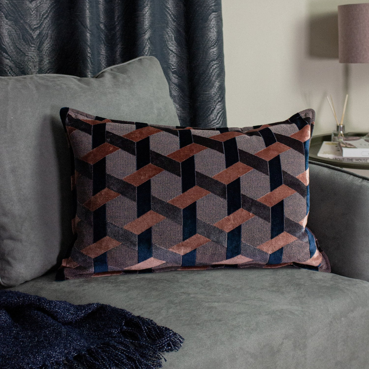 Delano Jacquard Velvet Cushion in blush, navy and charcoal, showing a woven geometric lattice pattern, styled on a grey sofa with a navy throw nearby.