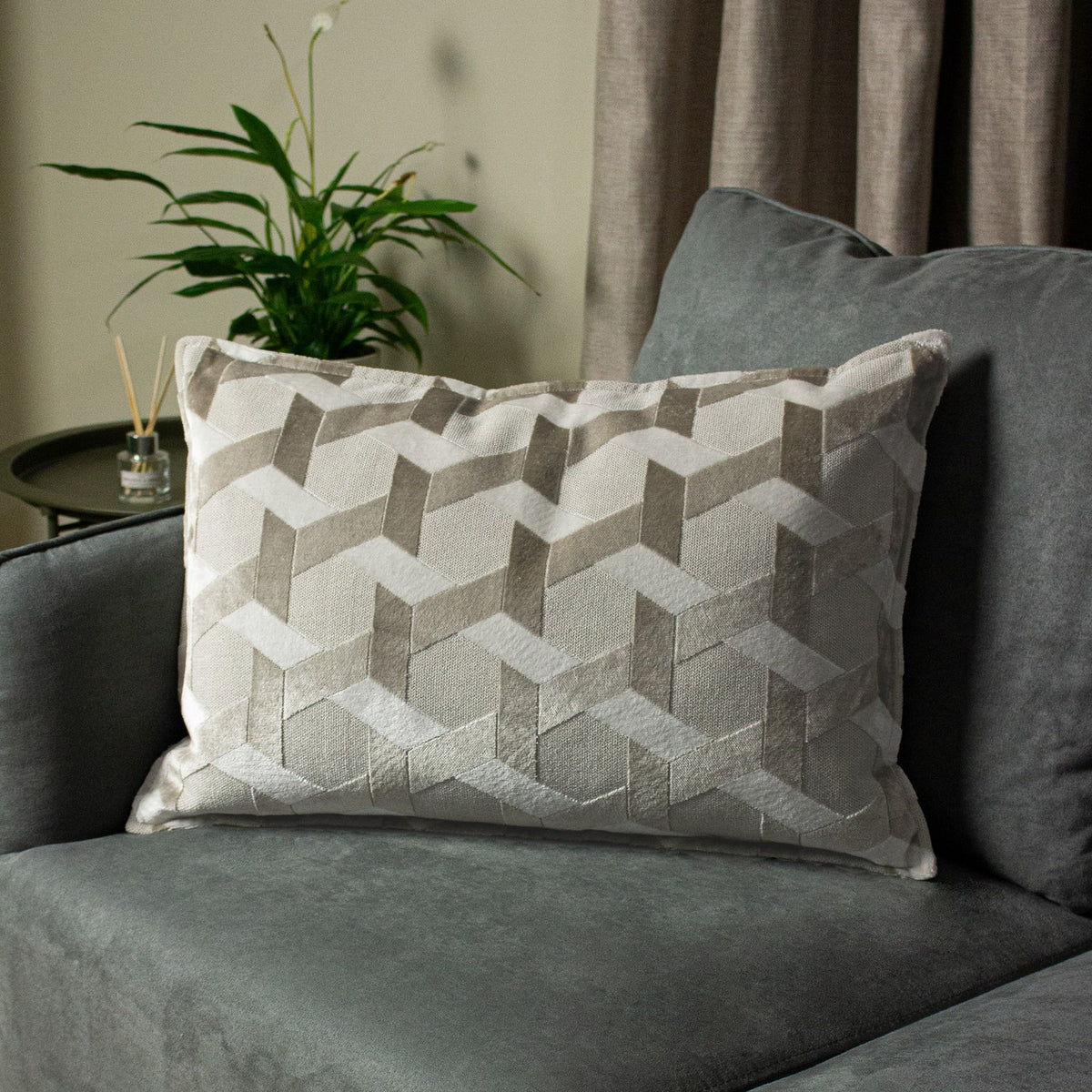 Delano Jacquard Velvet Cushion in ivory and taupe with a raised geometric chevron pattern, styled on a grey armchair beside a plant and side table.
