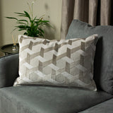 Delano Jacquard Velvet Cushion in ivory and taupe with a raised geometric chevron pattern, styled on a grey armchair beside a plant and side table.