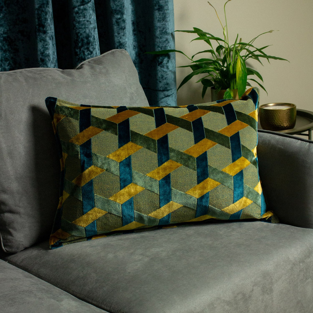Delano Jacquard Velvet Cushion in teal, gold and green with an interlocking lattice pattern, styled on a grey sofa near a teal curtain and plant.