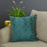 Delphi Velvet Linen Cushion in teal with maze-like jacquard pattern, shown on a grey sofa beside a plant and gold candle holder.