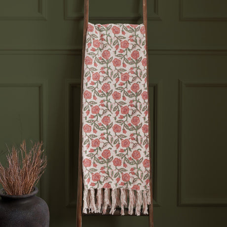 Delphine Floral Cotton Throw in coral pink and green on a cream cotton ground, with fringed edges, draped over a wooden ladder against a dark green panelled wall.