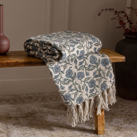 Delphine Floral Cotton Throw in Delph Blue, folded on a wooden bench with tasselled fringe, showing a cream cotton ground with dusty blue and green floral print.