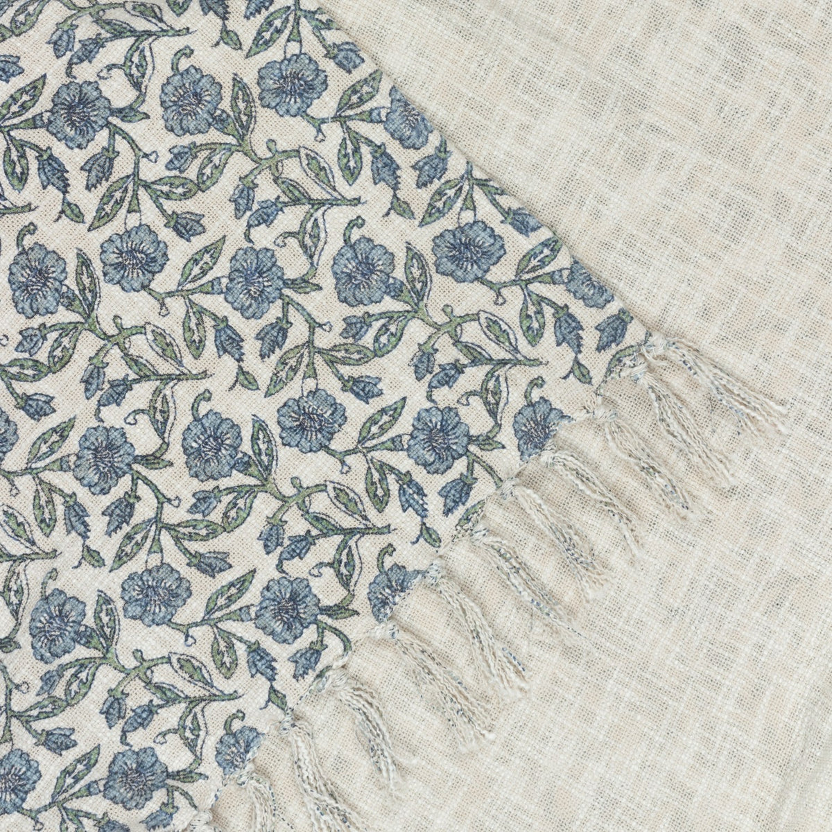 Delphine Floral Cotton Throw in Delph Blue, showing a cream cotton weave printed with dusty blue and green flowers, edged with knotted fringe.