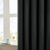 Black velvet eyelet curtain with soft matt pile, shown draped in folds beside a white-framed window with a mug and open book on the sill.
