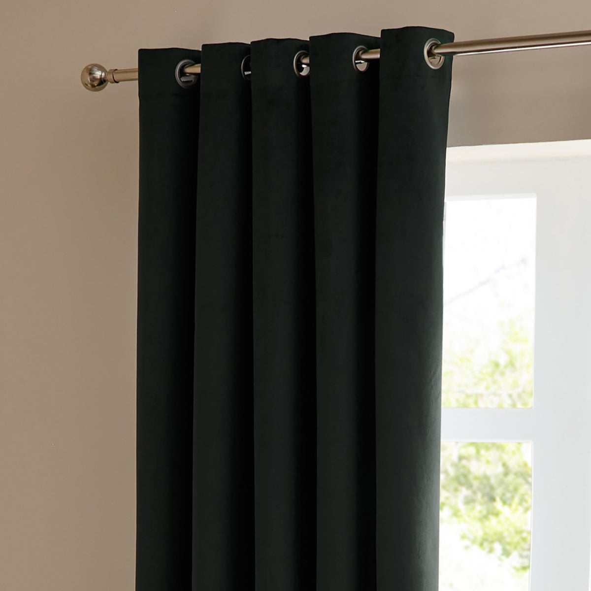 Velvet eyelet curtain in dark green-black, shown hanging in folds from a chrome pole with metal eyelet rings, in front of a window with daylight streaming in.