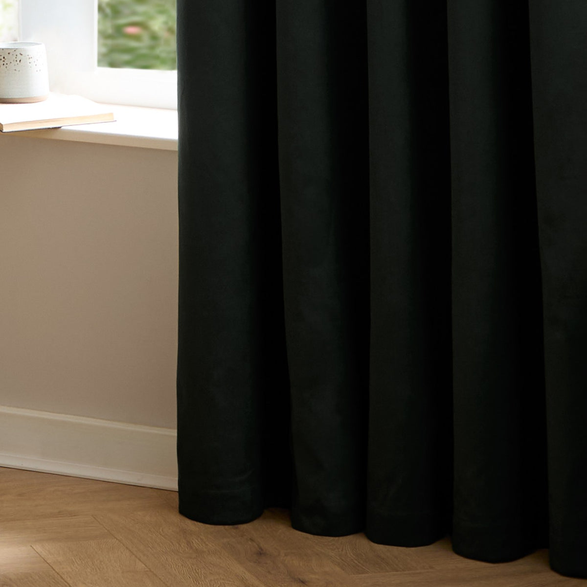 Black velvet eyelet curtain in a soft matt pile, shown falling in deep folds beside a window with a light wall and wooden herringbone floor.