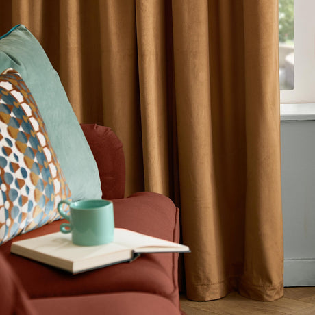 Caramel velvet eyelet curtains hanging in soft folds beside an armchair with teal and patterned cushions, shown in a living room setting.