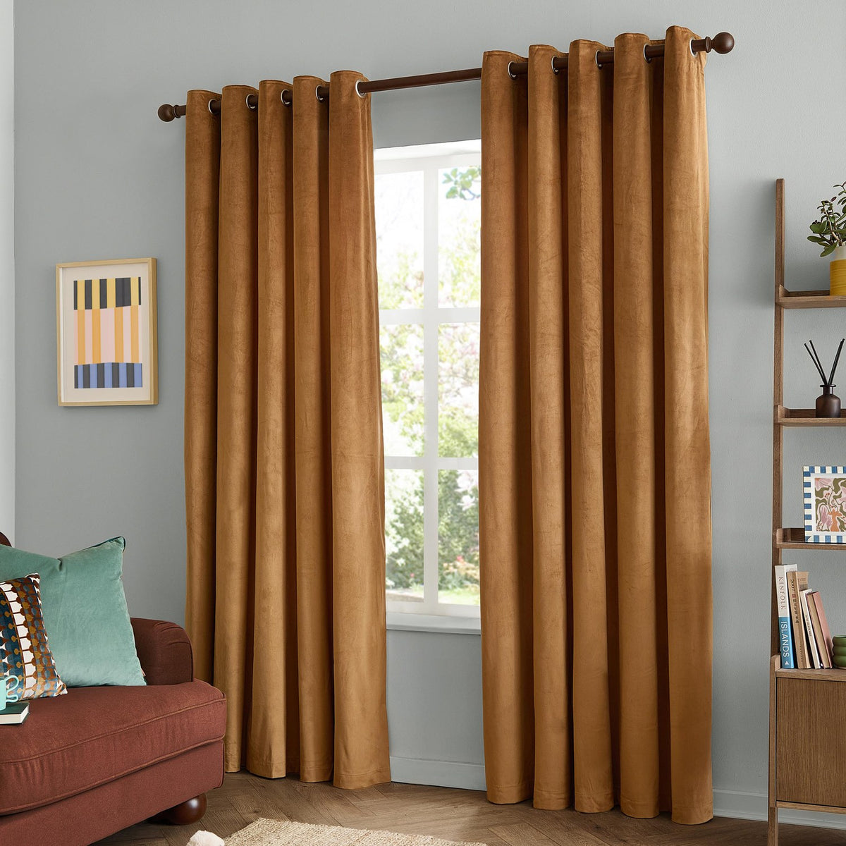 Caramel velvet eyelet curtains with brown grommets hang floor-length beside a window in a living room styled with a rust sofa and teal cushions.