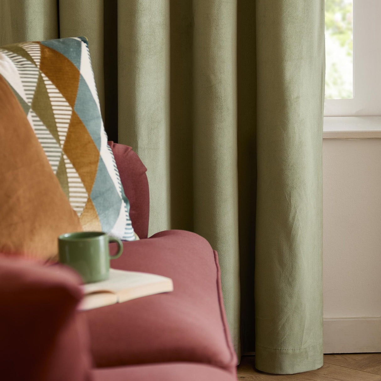 Green velvet eyelet curtain in soft matte fabric, shown draped in a living room beside a window with a pink armchair, patterned cushion, and mug in the foreground.