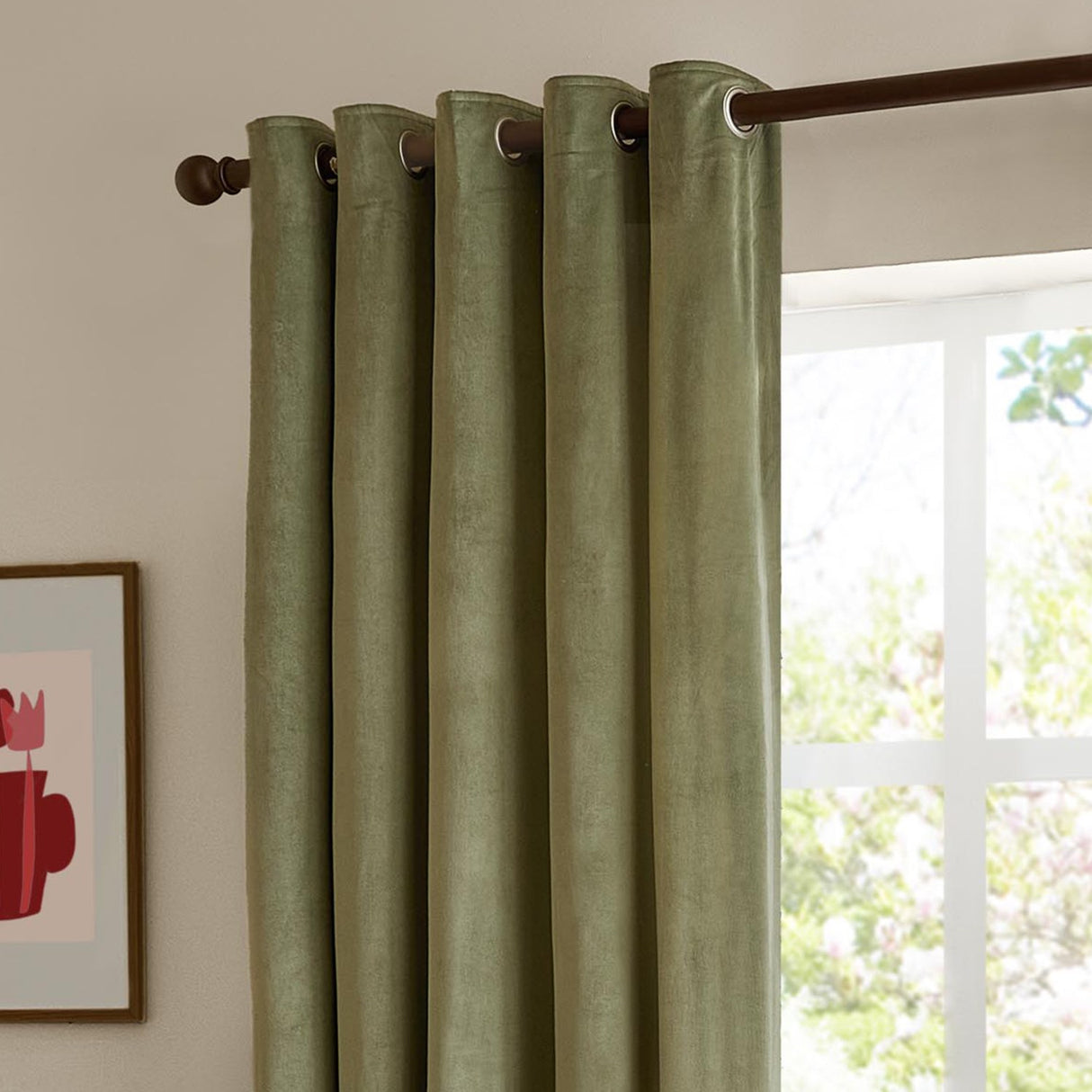 Green velvet eyelet curtains with metal grommets hung on a dark rod, shown in a living room beside a bright window and framed wall art.