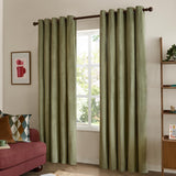 Green velvet eyelet curtains with grommet headings hung on a wooden pole, shown fully drawn in a living room beside a window, sofa, and shelving unit.