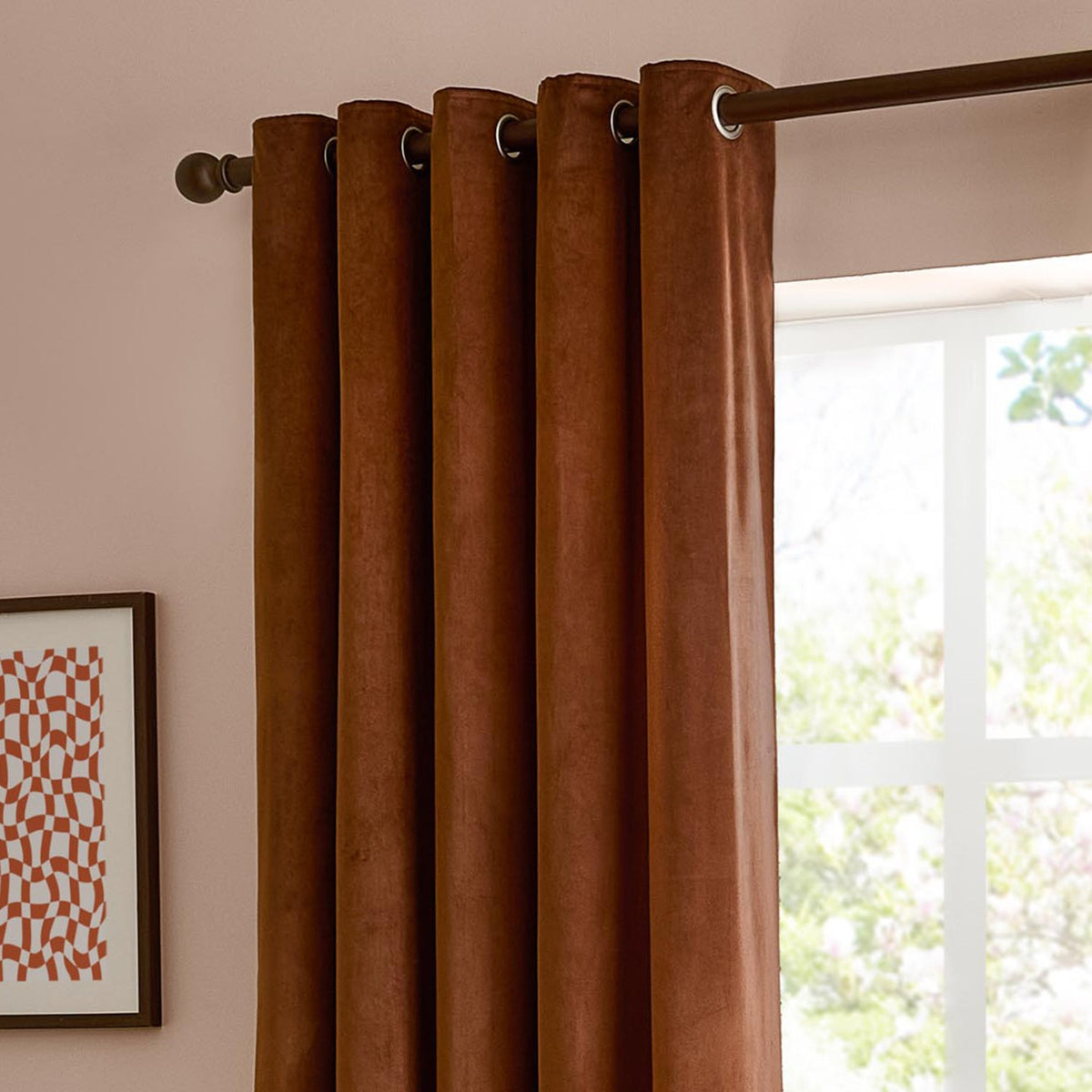 Velvet eyelet curtains in rust brown (Mink) hanging from a bronze pole on a pink wall beside a window, showing the plain matt pile and metal eyelet rings.