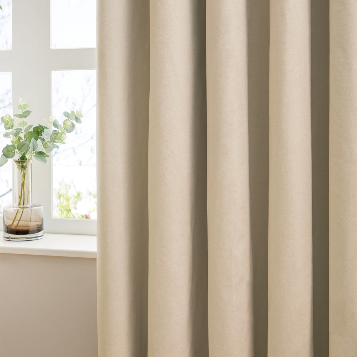 Close-up of The Ultimate Velvet Plain Lined Eyelet Curtains in natural beige, showing draped folds beside a window with a glass vase and eucalyptus stems.