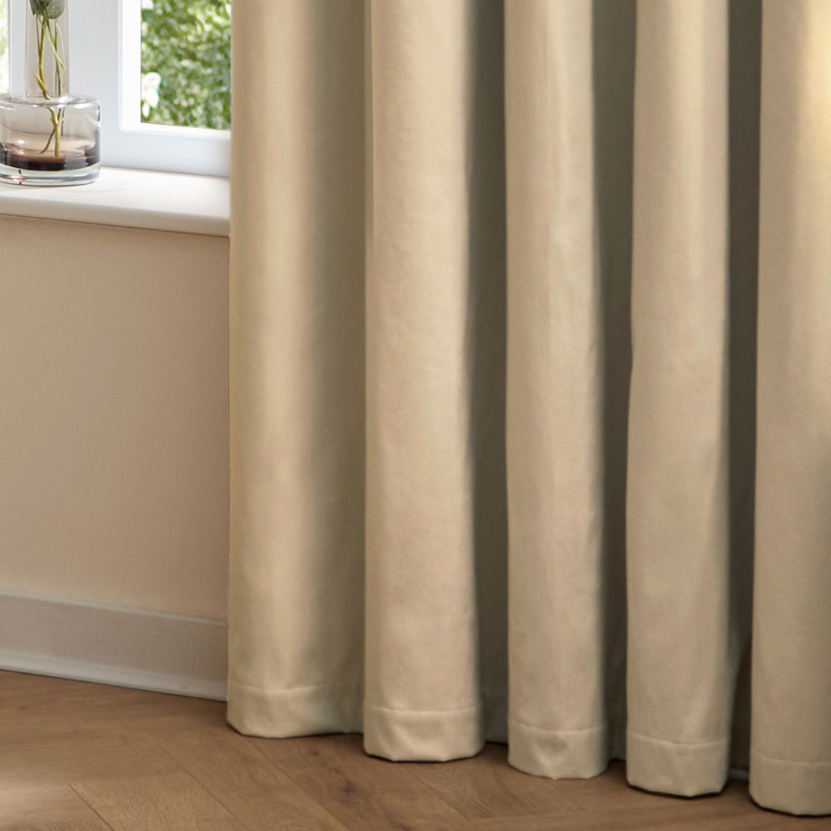 The Ultimate Velvet Plain Lined Eyelet Curtains in Natural, shown in soft folds pooling on a wooden floor beside a window in a bedroom setting.