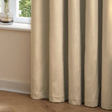 The Ultimate Velvet Plain Lined Eyelet Curtains in Natural, shown in soft folds pooling on a wooden floor beside a window in a bedroom setting.