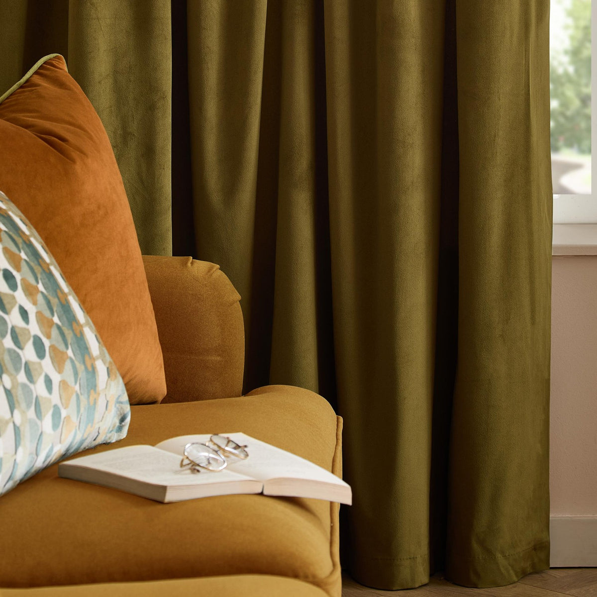 Olive green velvet eyelet curtains with soft folds, fully lined, shown draped by a window in a living room with mustard sofa and cushions.