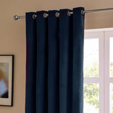 Royal blue velvet eyelet curtains hanging from a chrome pole beside a window, showing the matt fabric falling into rounded folds in a bedroom setting.
