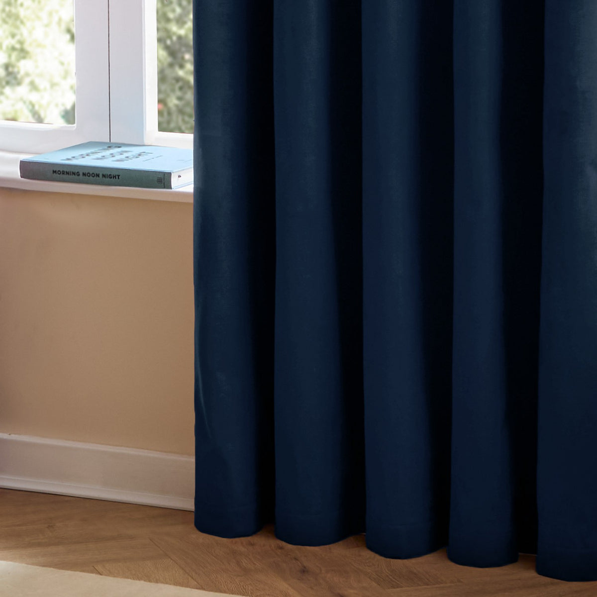 The Ultimate Velvet Plain Lined Eyelet Curtains in Royal blue, shown in soft folds by a window with a matt velvet finish, set beside a book on the sill and wooden flooring.