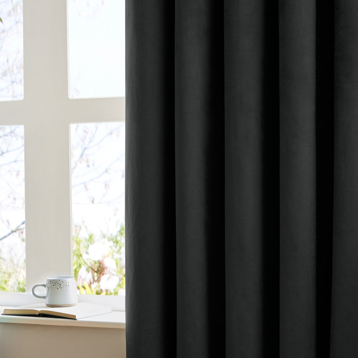 Black velvet pencil pleat curtain in soft vertical folds, shown beside a bright window with a mug and open book on the sill.