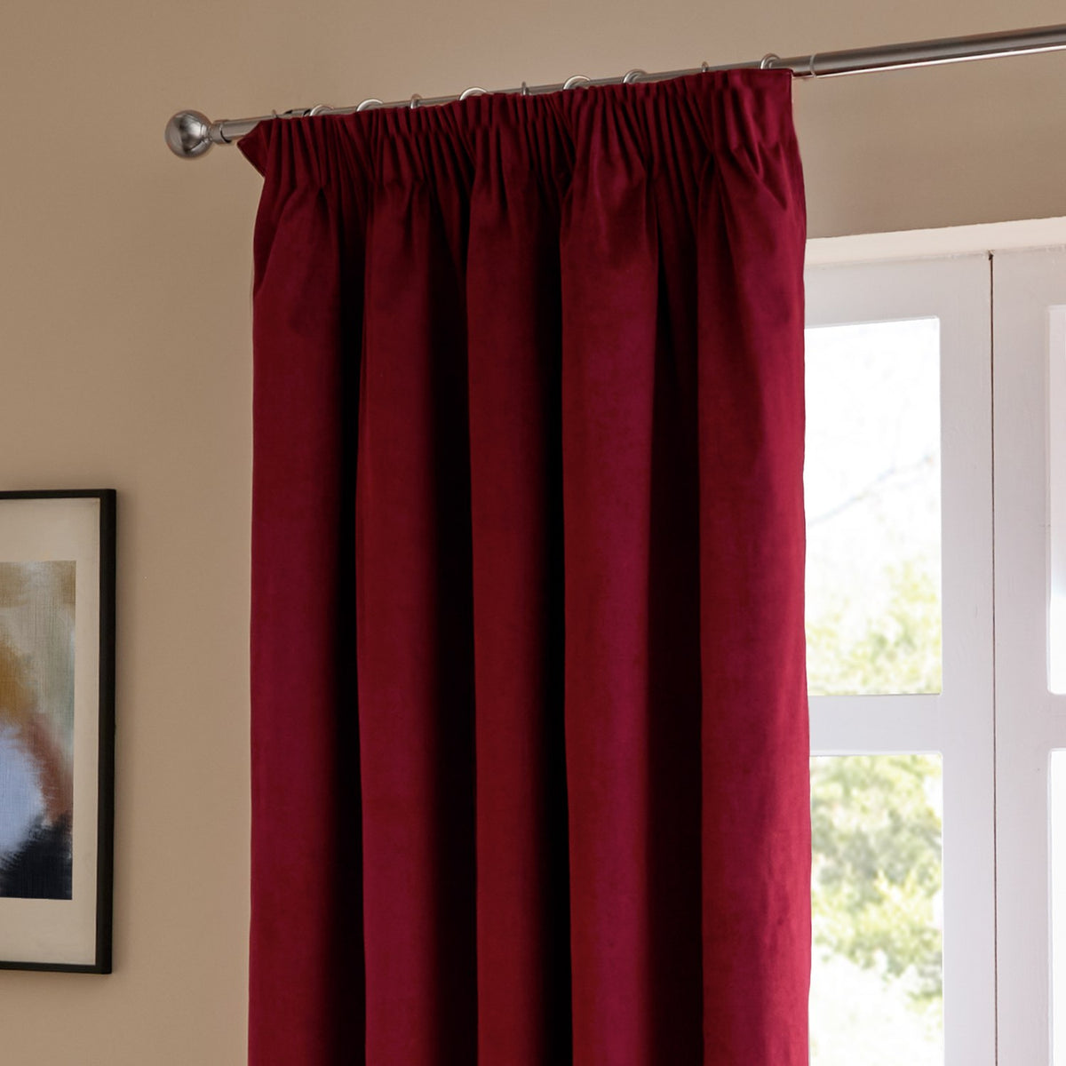 Burgundy velvet pencil pleat curtains with pencil-pleat heading on a chrome pole, shown hanging beside a window in a living room setting.