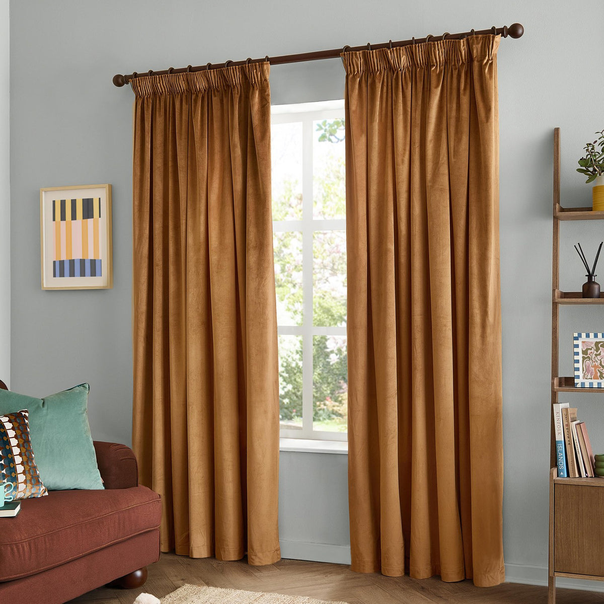 Caramel velvet pencil pleat curtains with pinch-pleat heading on a dark rod, shown as a pair framing a window in a living room with grey walls.