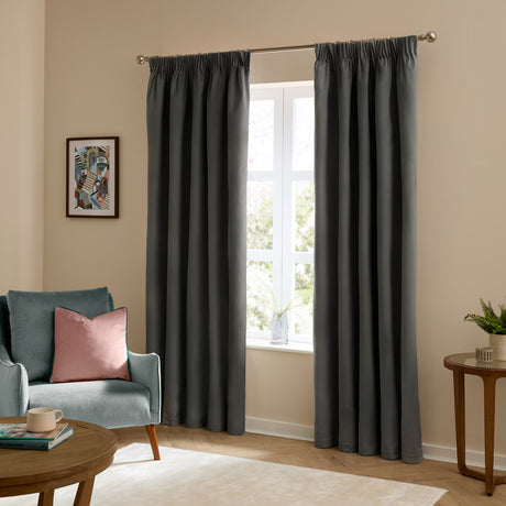Charcoal grey velvet pencil pleat curtains hanging on a metal pole either side of a window in a living room with a teal armchair and pink cushion nearby.
