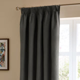 Charcoal grey velvet pencil pleat curtain hanging from a chrome rod on rings, shown against a beige wall beside a bright window in a living room.