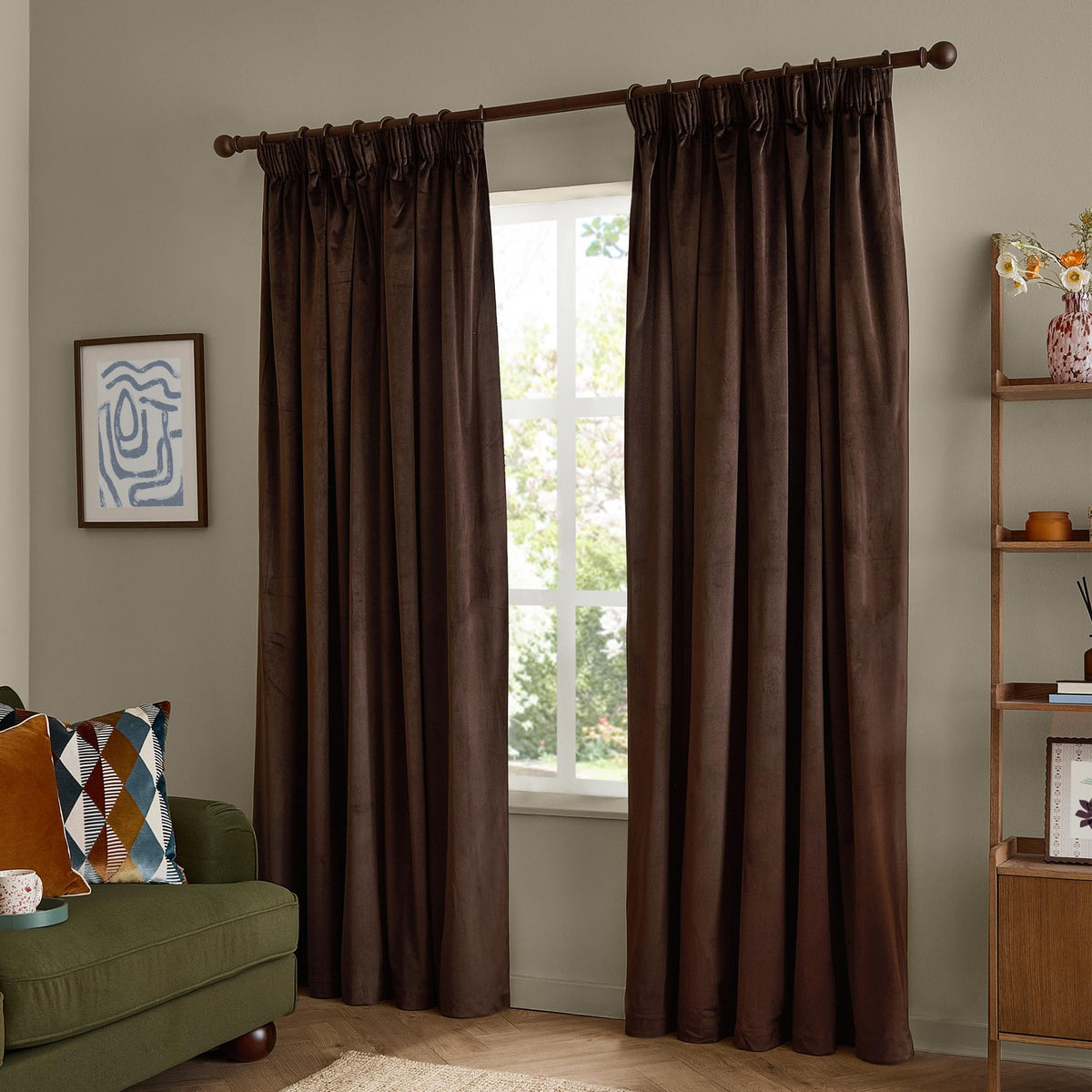 Chocolate brown velvet pencil pleat curtains with pinch-pleat heading on a wooden pole, shown framing a window in a living room with a green sofa and ladder shelf.