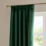 Emerald green velvet pencil pleat curtain hung on a brass rod with rings, showing gathered header folds beside a bright window.
