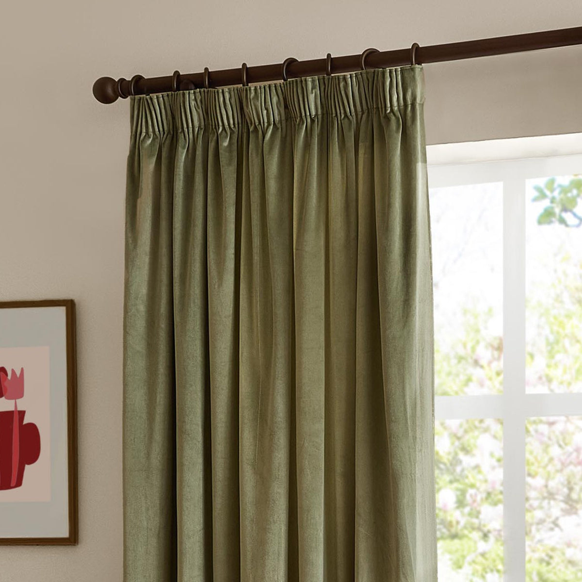 Green velvet pencil pleat curtains with pinch-pleated heading on a dark rod, hung beside a bright window in a living room setting.