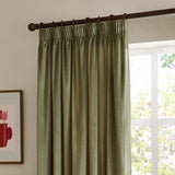 Green velvet pencil pleat curtains with pinch-pleated heading on a dark rod, hung beside a bright window in a living room setting.