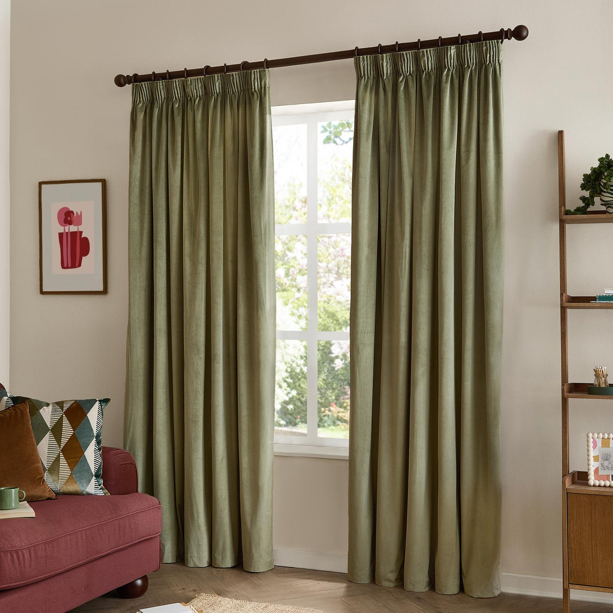Green velvet pencil pleat curtains with pinch-pleated heading on a dark pole, hung in a living room beside a window, framed print, and wooden ladder shelf.