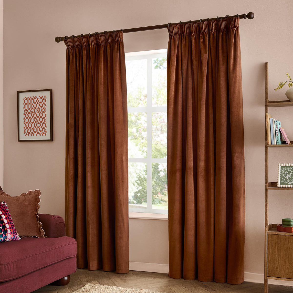 Rust velvet pencil pleat curtains with pinch-pleated heading on a dark pole, hanging in soft folds beside a window in a living room.