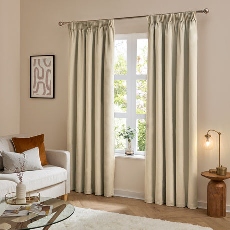 The Ultimate Velvet Polyester Pencil Pleat Curtains in natural beige hang from a chrome rod either side of a window in a living room with a cream sofa and armchair.