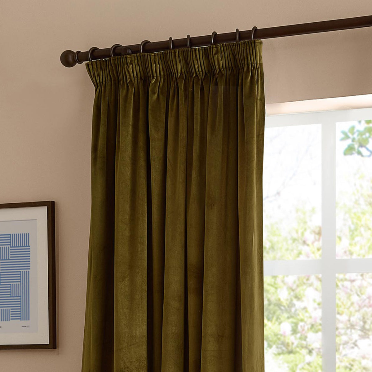Olive green velvet pencil pleat curtain hanging on a dark rod with pleated header tape and rings, shown beside a window in a living room setting.