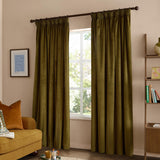 Olive green velvet pencil pleat curtains hanging on a dark rod in a living room, framed by a mustard sofa, bookshelf, and window with daylight.