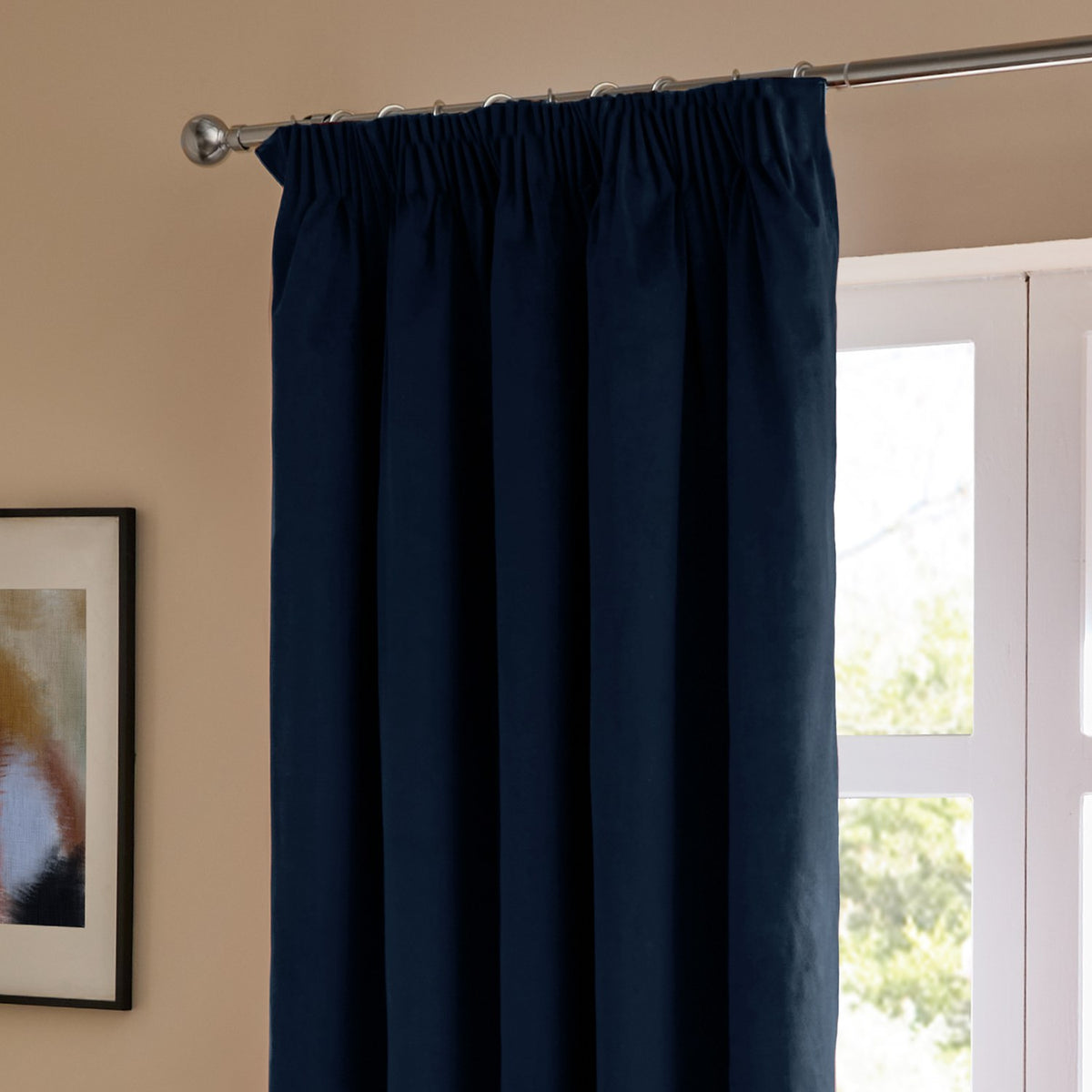 Royal blue velvet pencil pleat curtain with pinch-pleated header on a chrome pole, shown draped beside a window in a warm-toned living room.