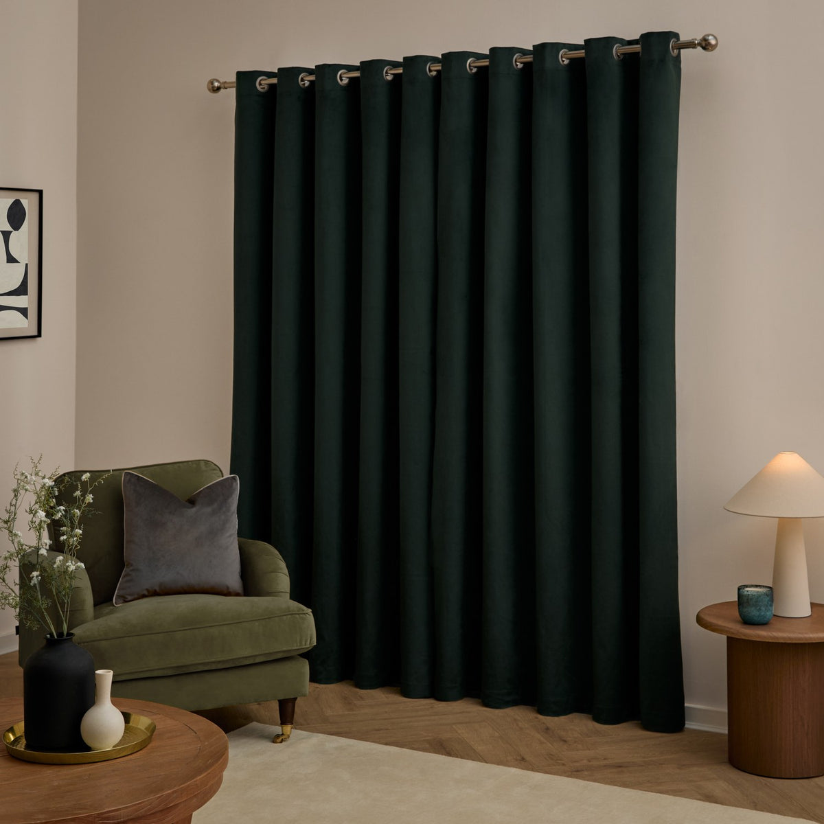 Dark green velvet eyelet curtains hung on a metal pole in a living room with a green armchair, wooden side tables, and a table lamp.