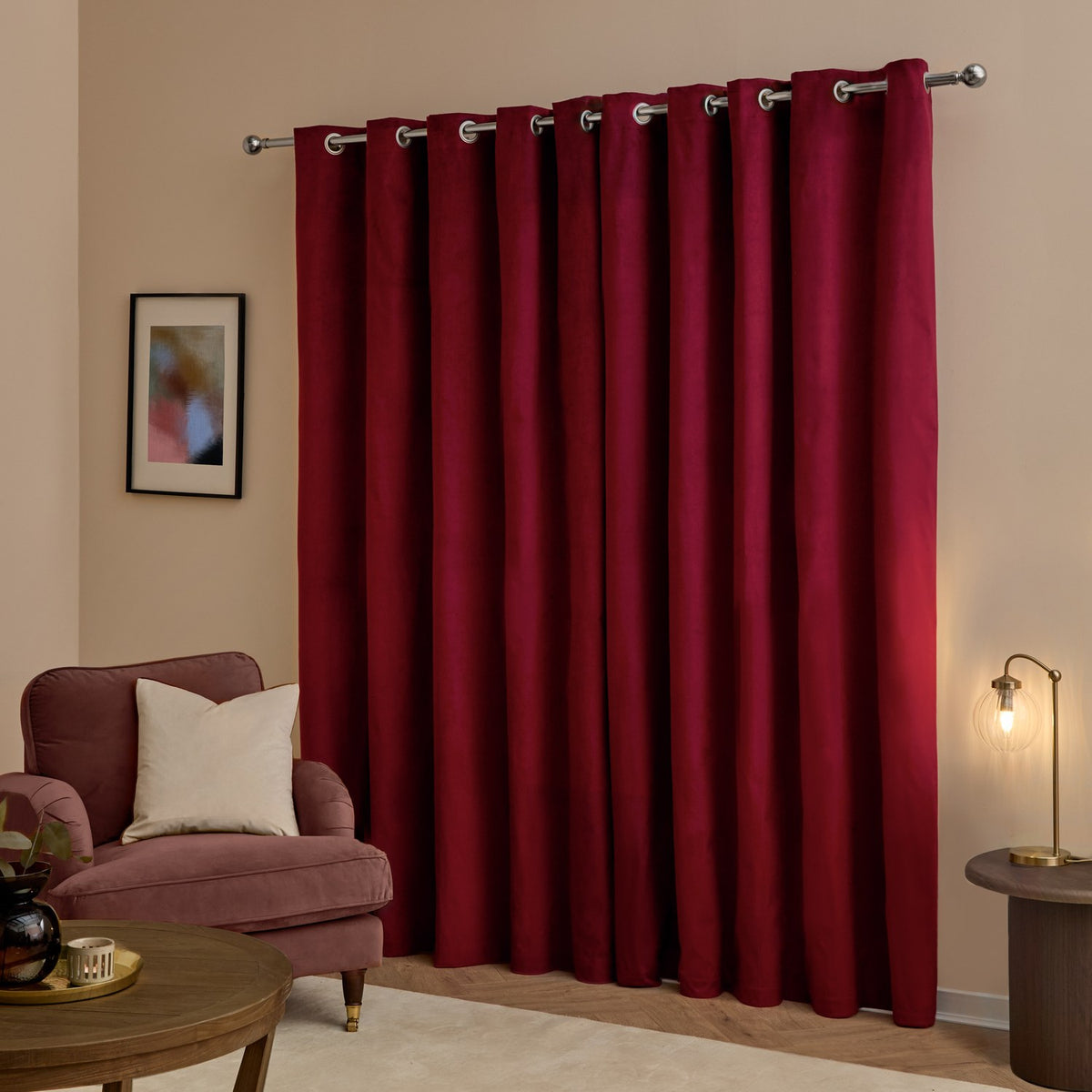 Burgundy velvet eyelet curtains with silver grommets hung on a chrome pole, shown floor-length in a living room beside an armchair and floor lamp.