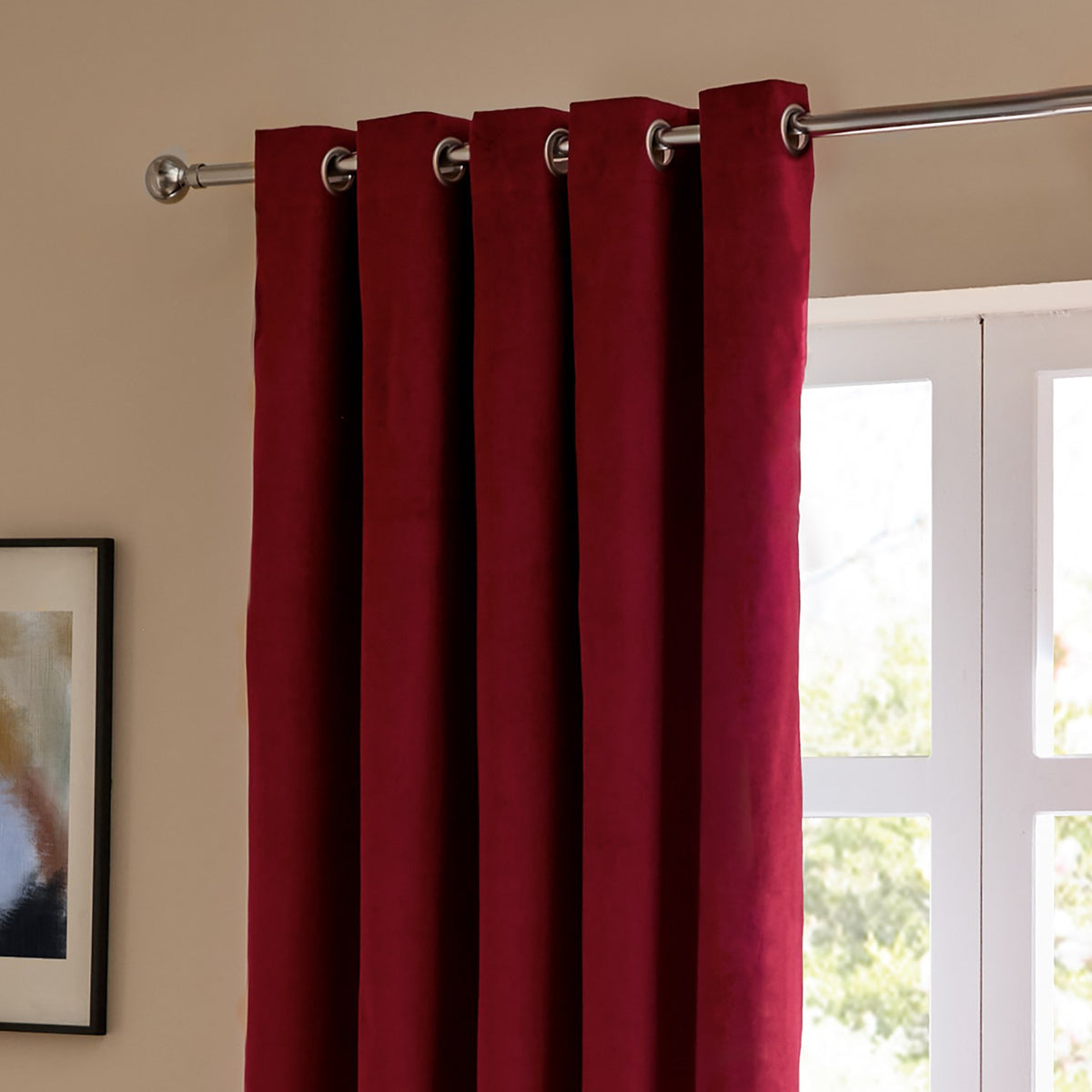 Burgundy velvet eyelet curtain hung on a chrome pole beside a window, showing matt fabric folds and metal grommets in a living room setting.