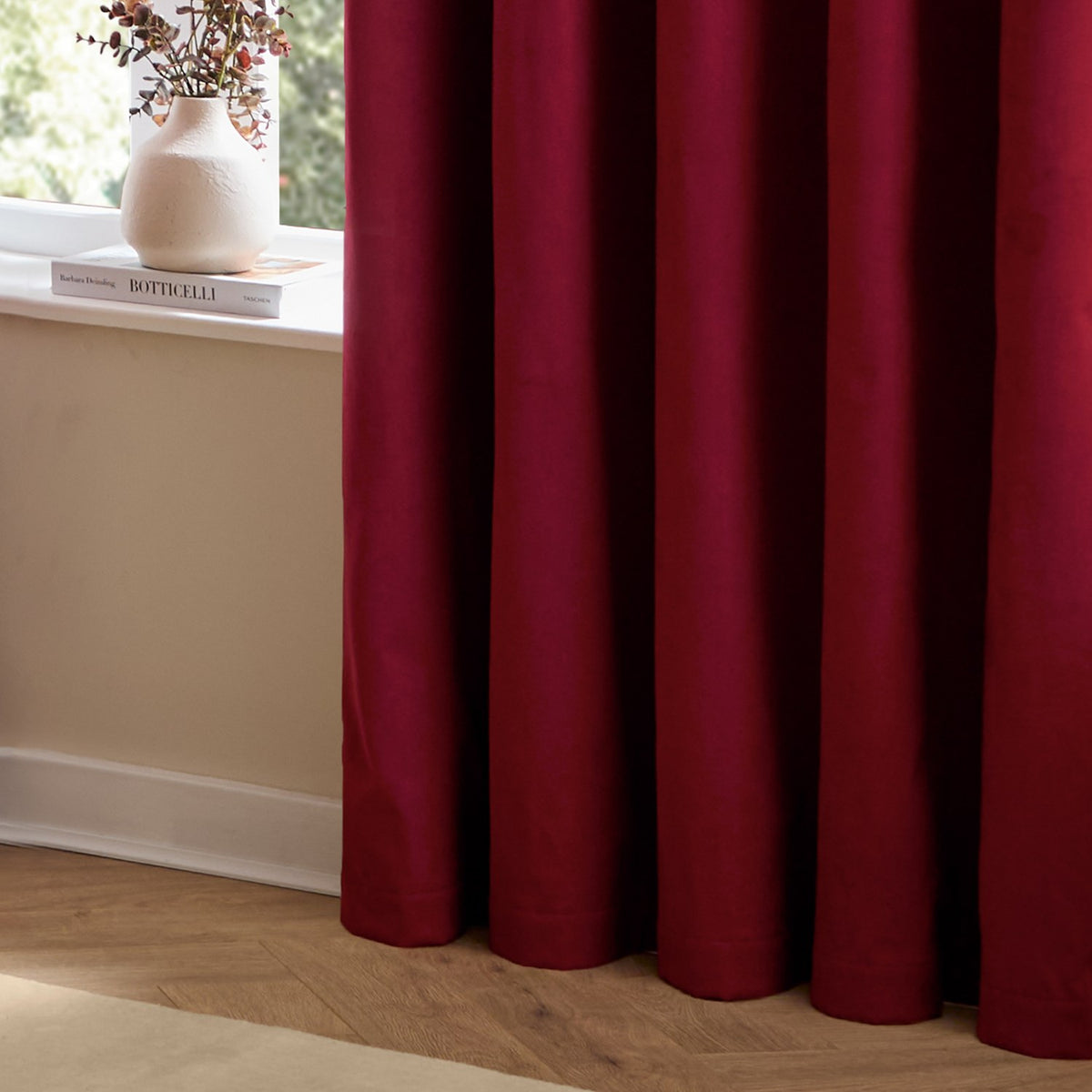 The Ultimate Velvet Plain Lined Eyelet Curtains in Burgundy shown in soft folds by a window with a beige wall, wooden floor, and vase on the sill.